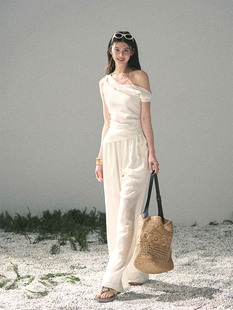 Woman wearing a white off-shoulder top and wide-leg pants with a woven beach tote