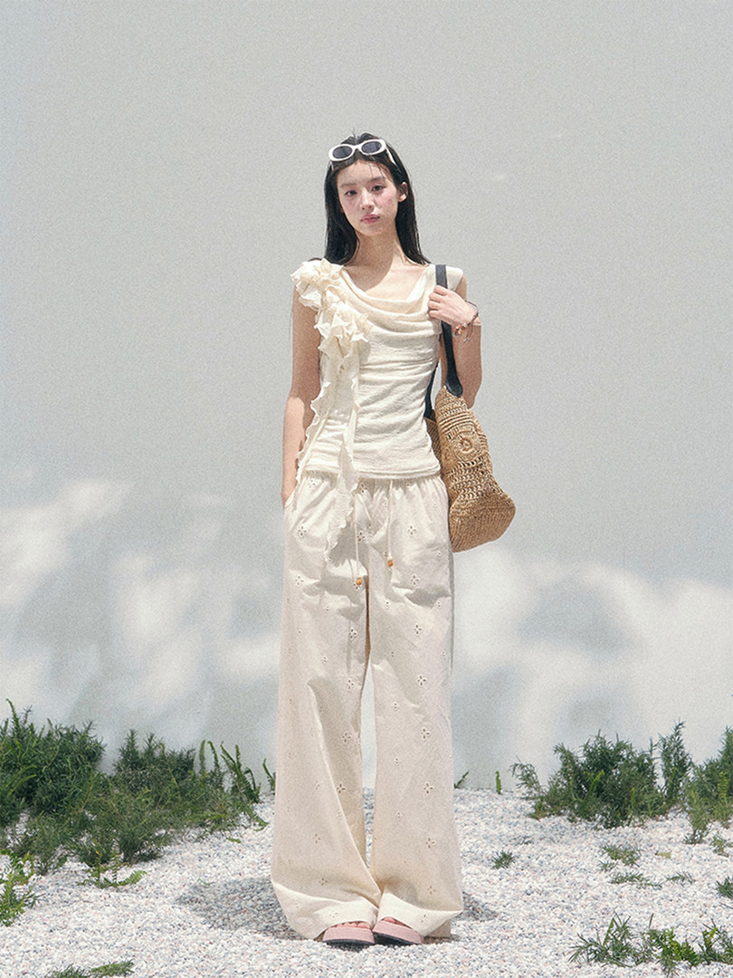 Model wearing a white ruffled top and wide-leg pants with a woven tote bag outdoors