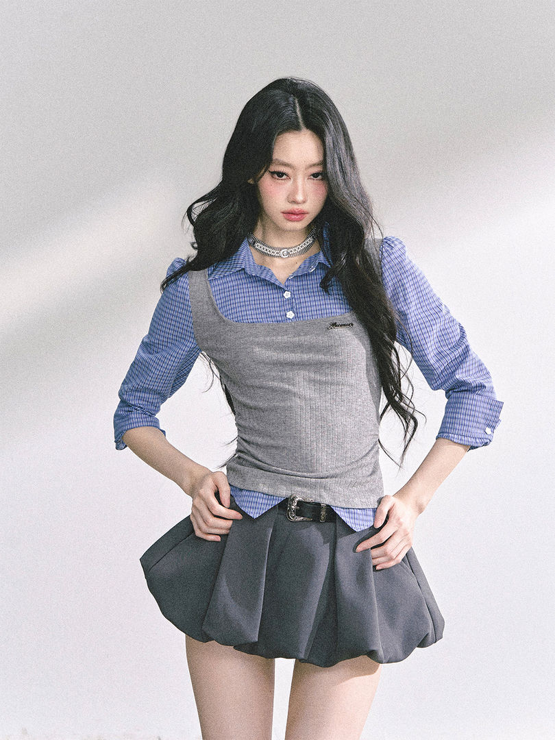 Model wearing a blue check shirt layered with a gray fitted camisole and dark pleated mini skirt