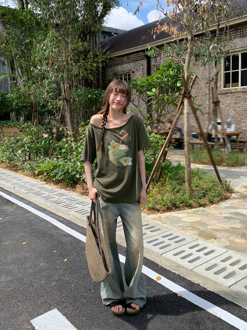 Model wearing an oversized olive graphic T-shirt, wide-leg jeans, and a woven tote bag outdoors