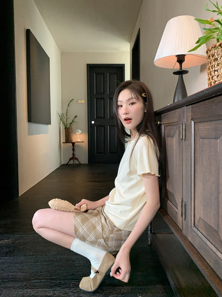 Woman modeling a cream top with plaid shorts and beige flats indoors