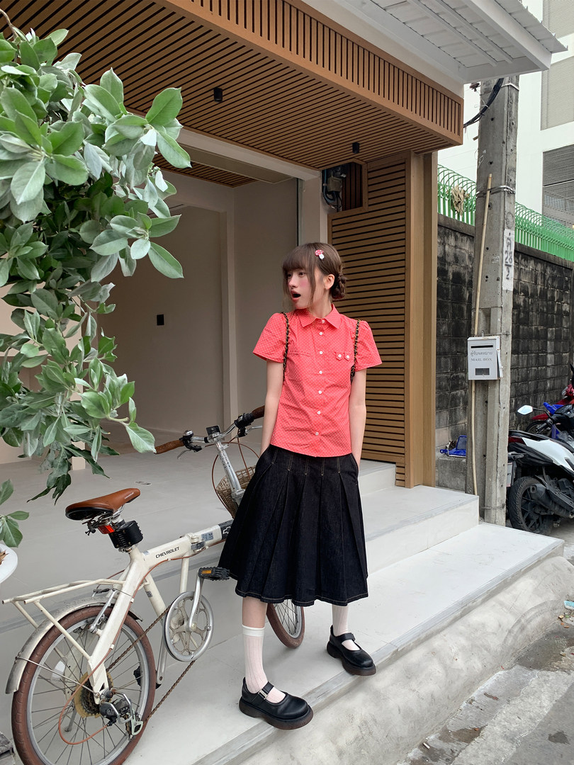 Young woman wearing a coral short-sleeve button-up top with a black pleated skirt and Mary Jane shoes