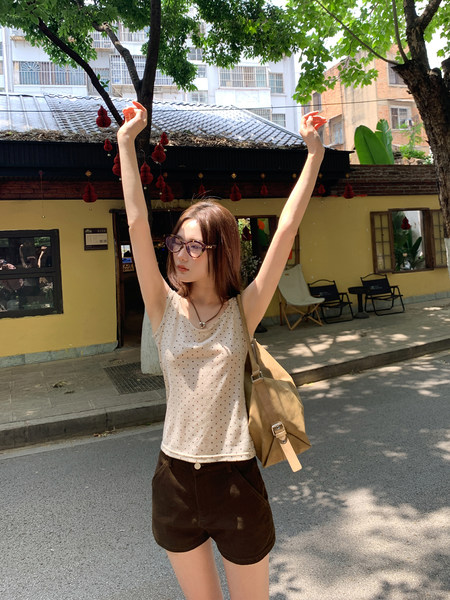Woman wearing a beige polka dot tank top with dark shorts and a tan shoulder bag outdoors