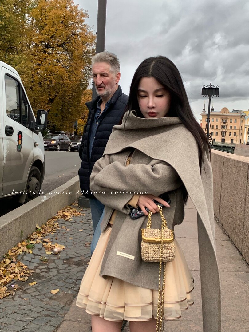Fashion model wearing a gray oversized coat with a cream skirt and small chain handbag outdoors