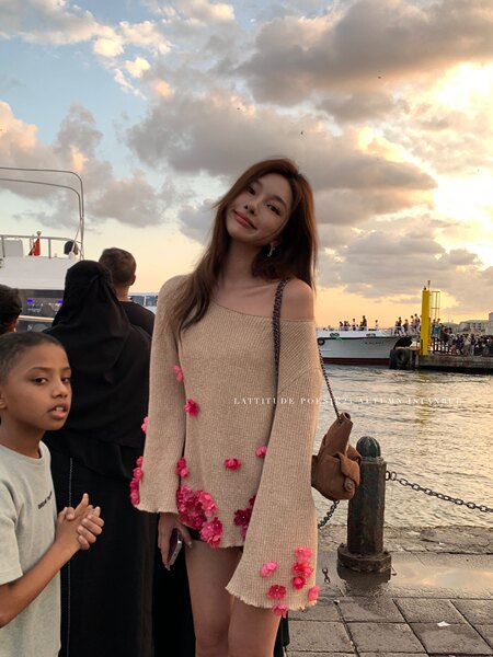 Woman wearing a beige off-shoulder knit dress with pink floral details by the waterfront at sunset