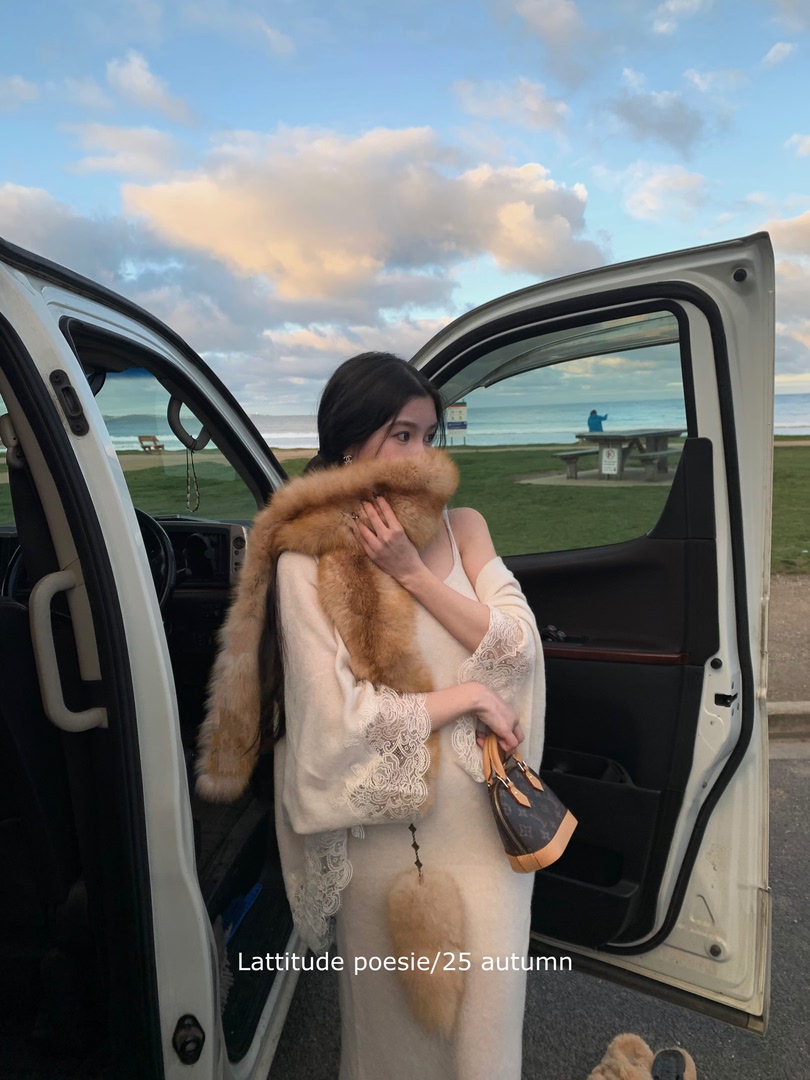 Woman wearing a cream lace dress with a fur stole and holding a small handbag by the beach