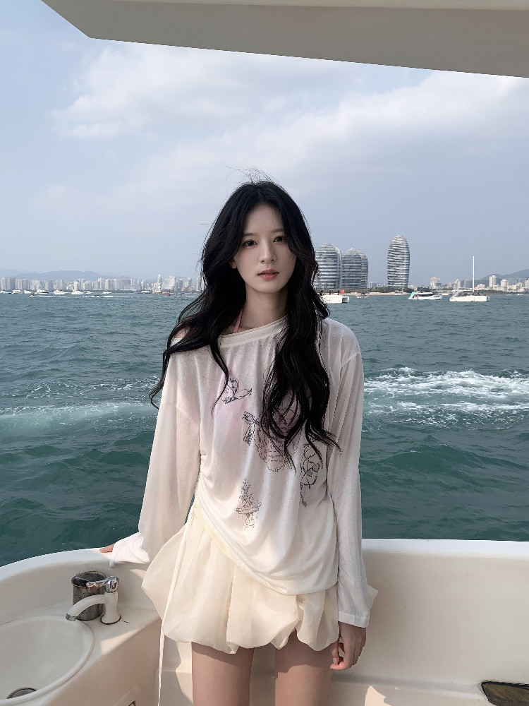 Model wearing a sheer long-sleeve top with a light skirt on a boat by the sea