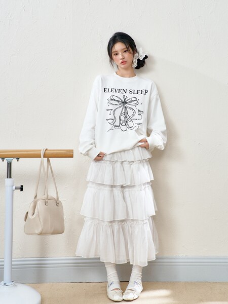 Model wearing a white graphic sweatshirt and layered white skirt with handbag and flats