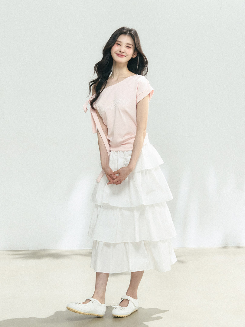 Model wearing a pale pink one-shoulder top with a white tiered midi skirt and white shoes