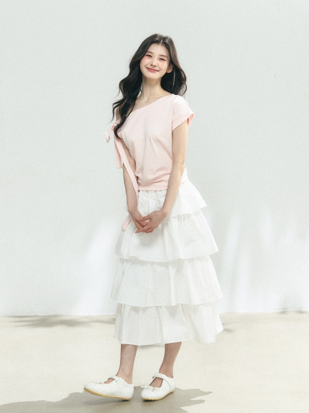 Model wearing a pale pink one-shoulder top with a white tiered midi skirt and white shoes
