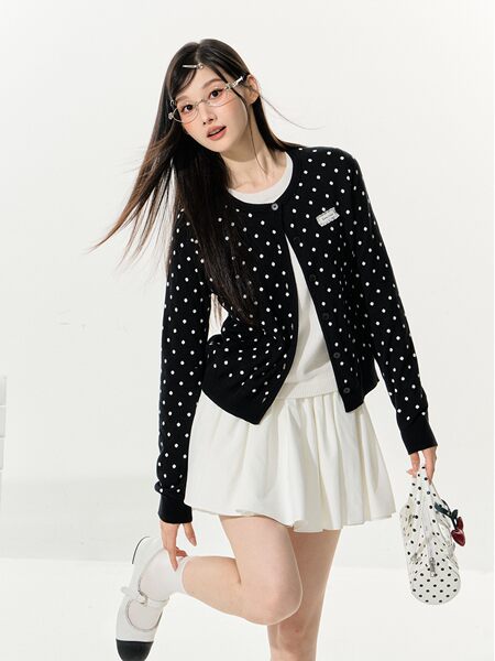 Model wearing a black polka dot cardigan with a white pleated skirt and white shoes