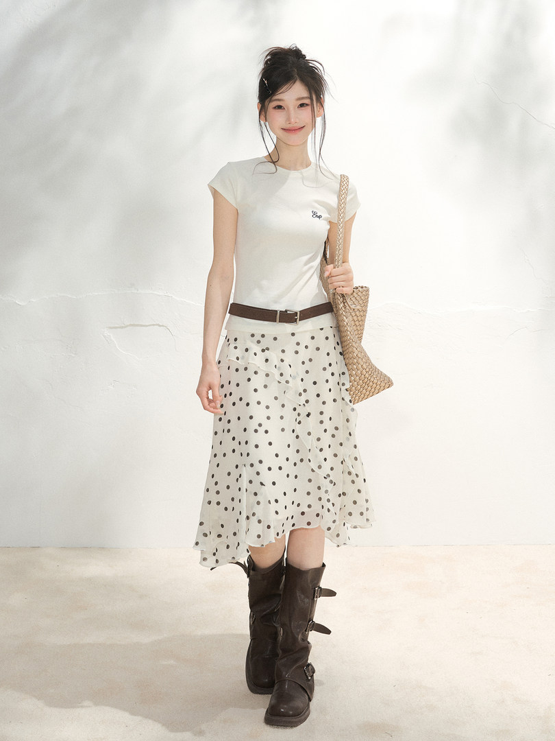 Model wearing a white top with a polka dot skirt, brown belt, woven bag, and tall boots