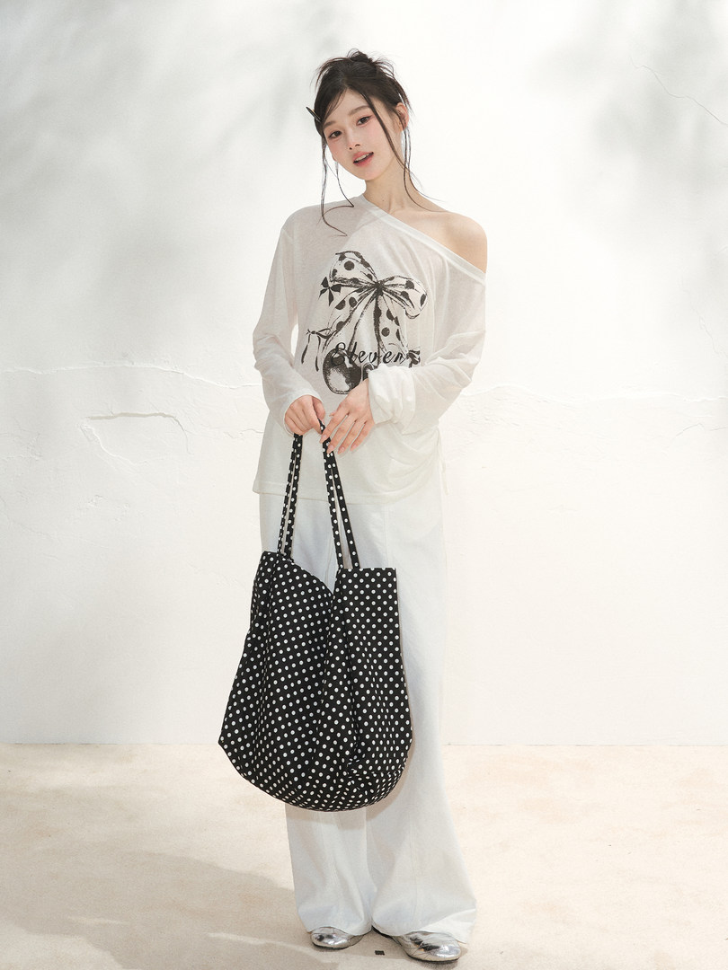 Model wearing a white off-shoulder graphic top with loose white pants and a black polka dot tote bag