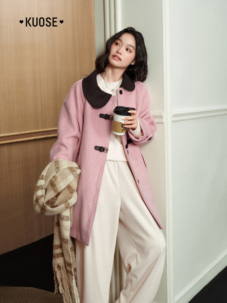Model wearing a pink coat with a dark collar and cream pants, holding a coffee cup