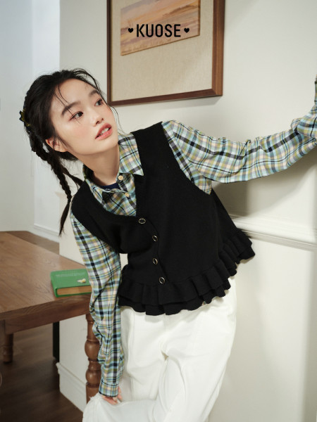 Model wearing a black ruffle vest over a plaid shirt with white pants