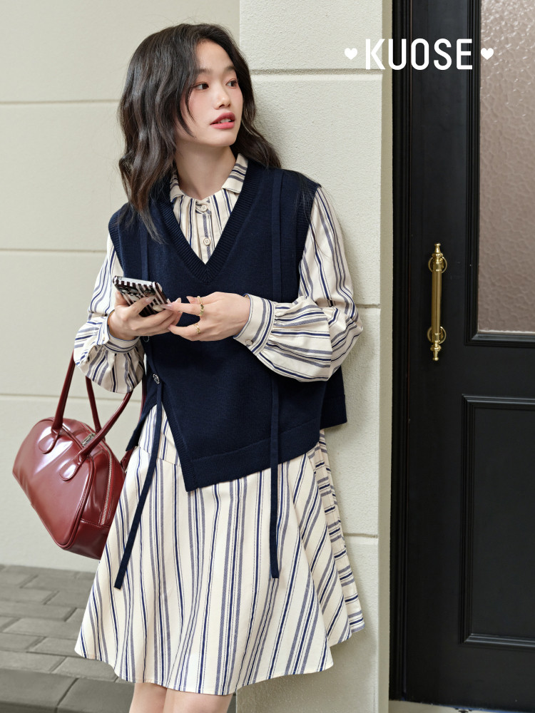 Model wearing a striped shirt dress layered with a navy sweater vest and carrying a red handbag