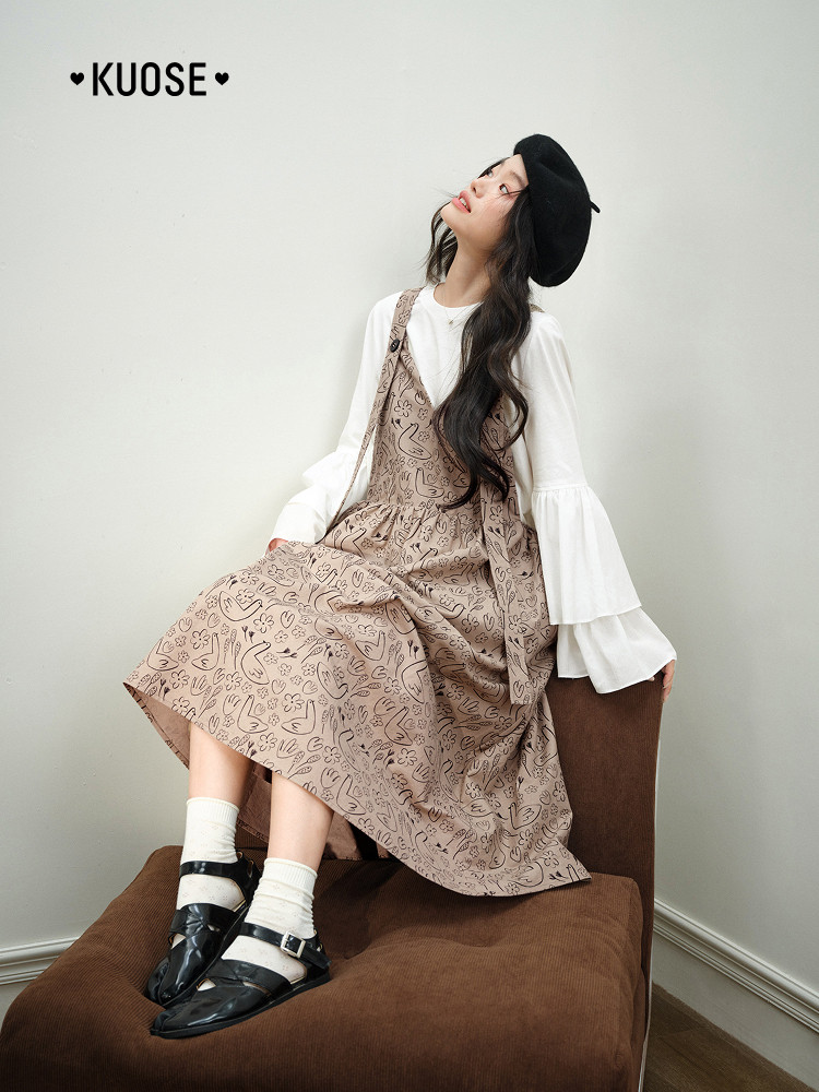 Model wearing a beige patterned pinafore dress over a white blouse with black shoes and beret