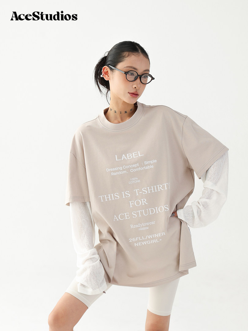 Model wearing a beige oversized graphic T-shirt layered over a white long-sleeve top