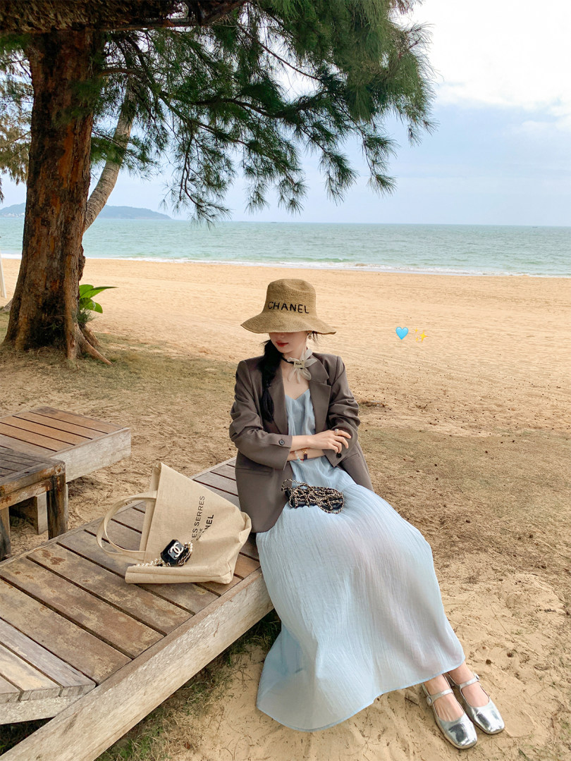 Woman wearing a light blue dress and blazer with a straw hat and handbag on a beach