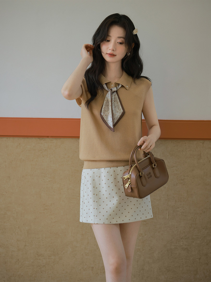 Model wearing a beige sleeveless knit top with a scarf tie and a white polka dot skirt, holding a brown handbag