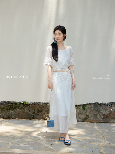 Model wearing a white two-piece summer outfit with a sheer long skirt and blue sandals