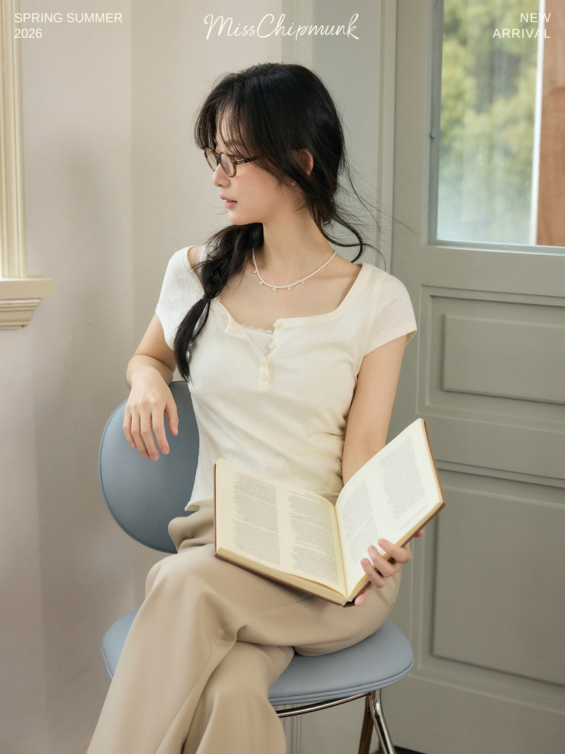 Model wearing a cream short-sleeve top and beige pants, seated indoors with a book