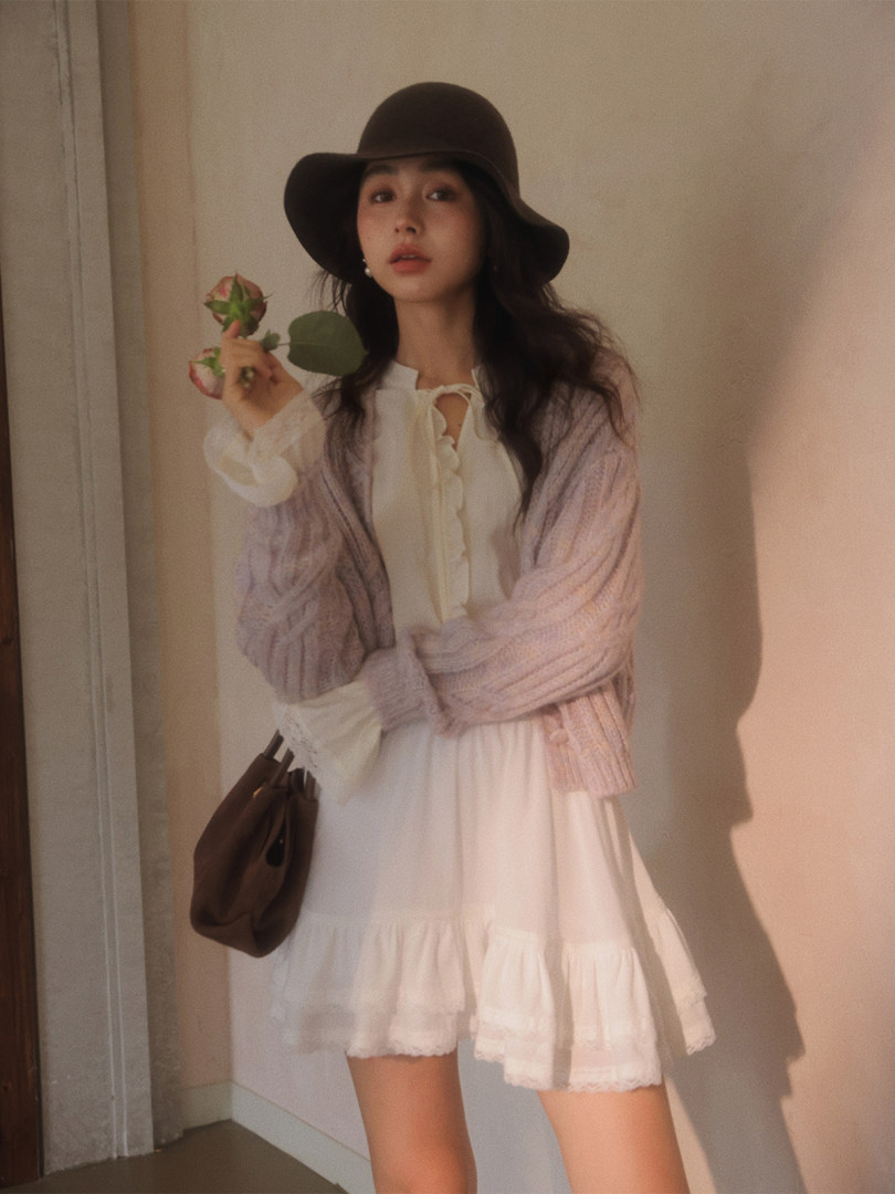 Woman wearing a white dress with a pastel cardigan, dark hat, and brown handbag