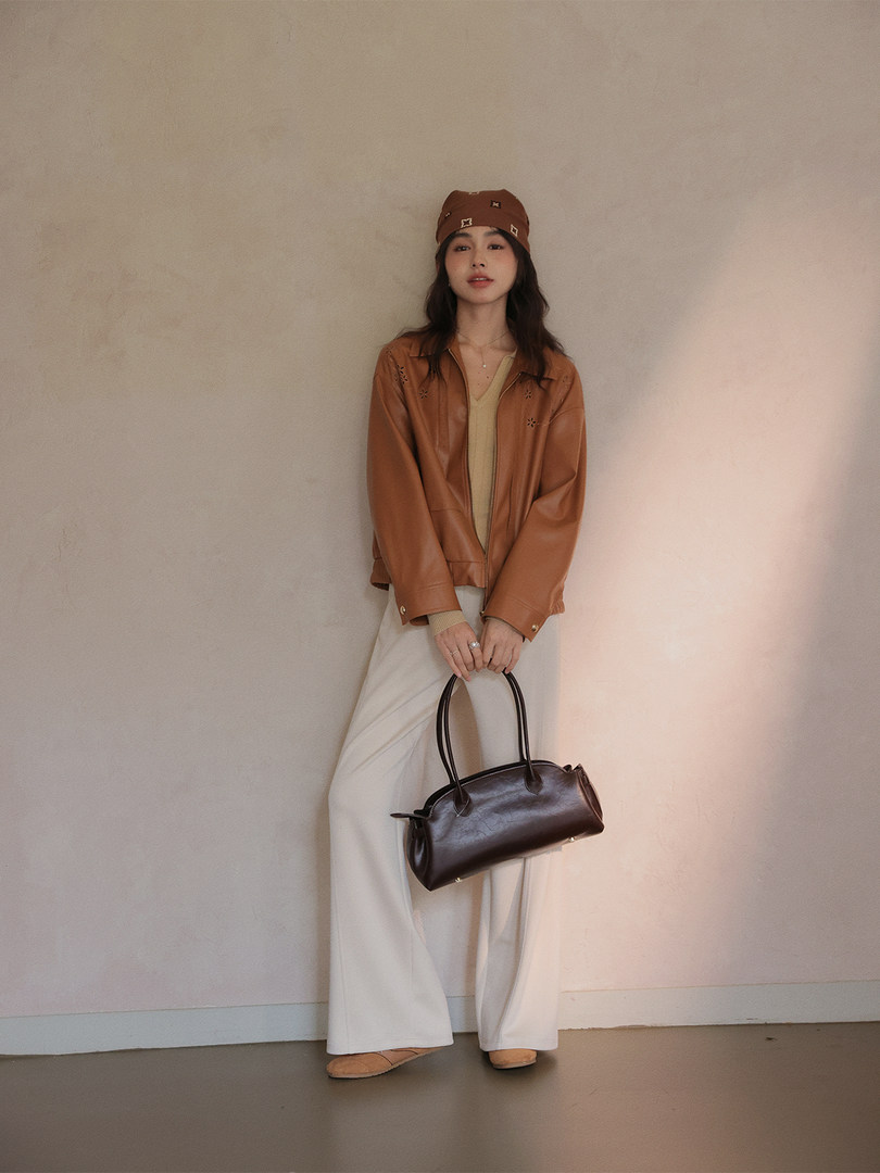 Model wearing a brown jacket with cream wide-leg pants and holding a dark handbag