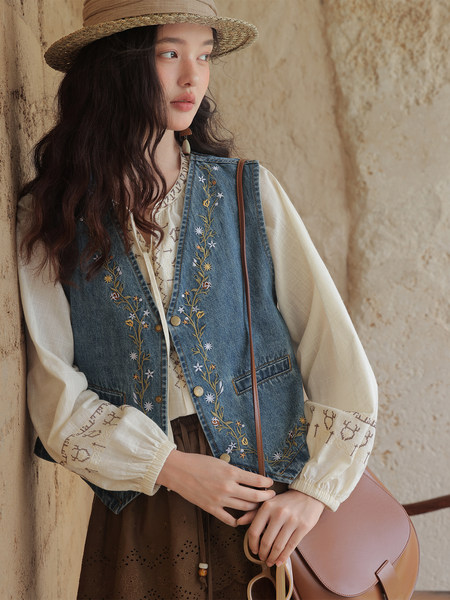 Model wearing an embroidered denim vest over a blouse with a straw hat and crossbody bag