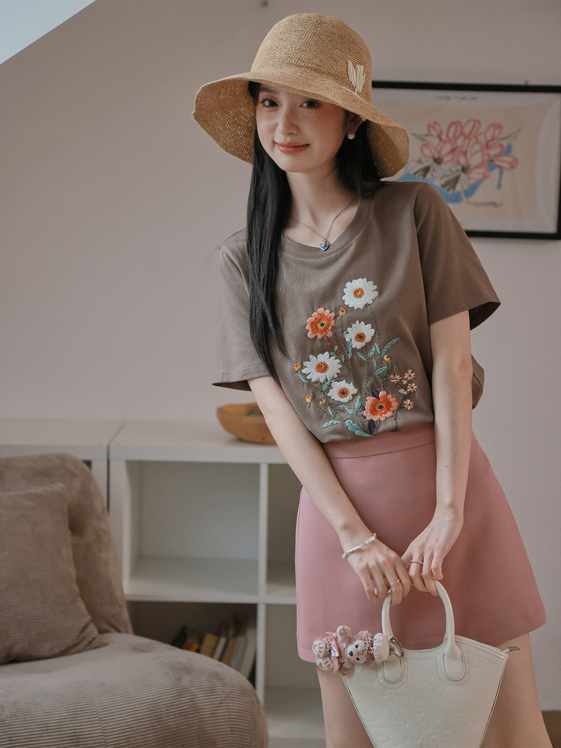 Woman modeling a brown embroidered floral top with pink skirt and straw hat, holding a white handbag