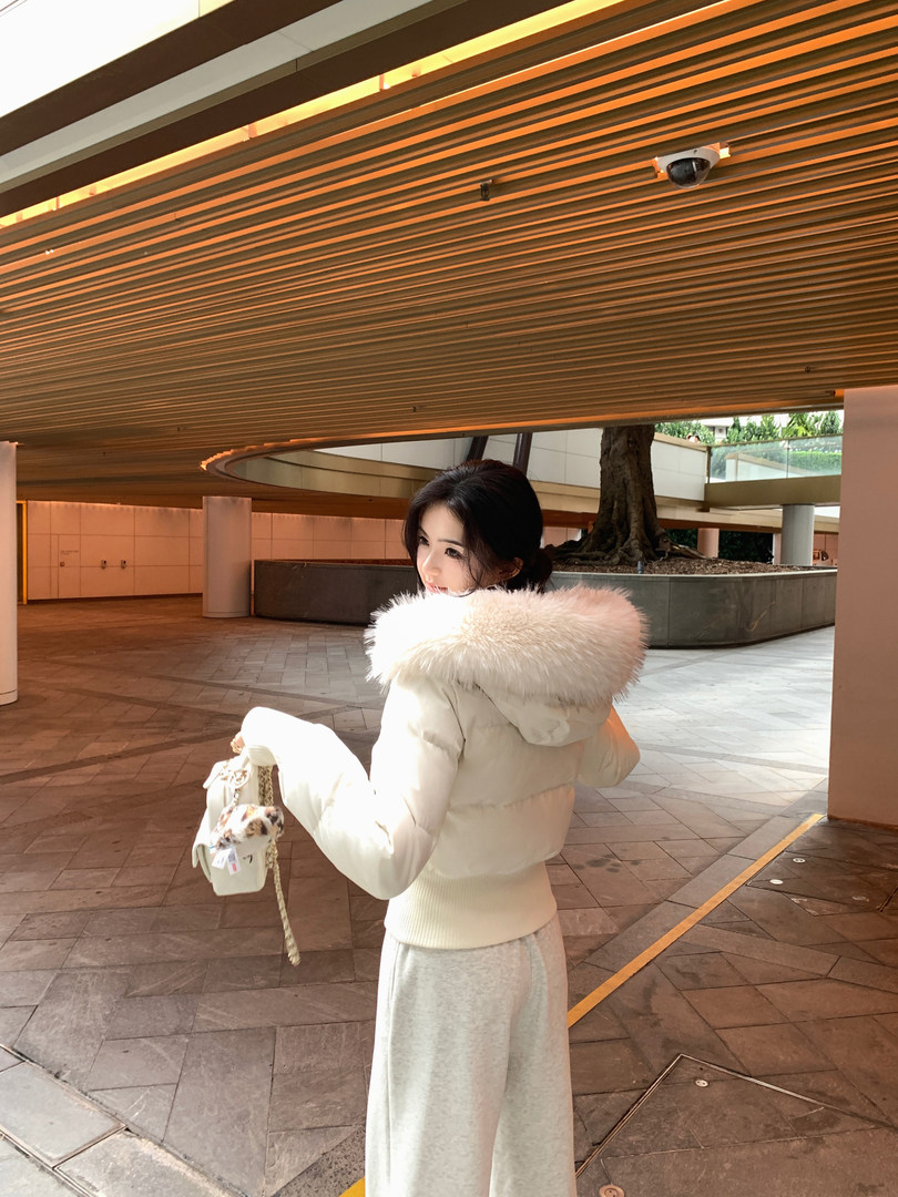 Woman wearing a white puffer jacket with a fur-lined hood, paired with gray pants and holding a small handbag.