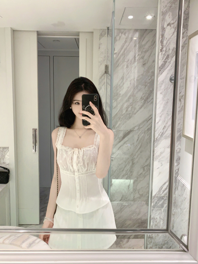 Woman taking mirror selfie wearing white lace top and skirt in bathroom