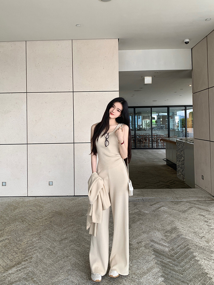 Woman wearing a beige sleeveless jumpsuit with wide legs, holding a matching jacket, and carrying a white handbag.