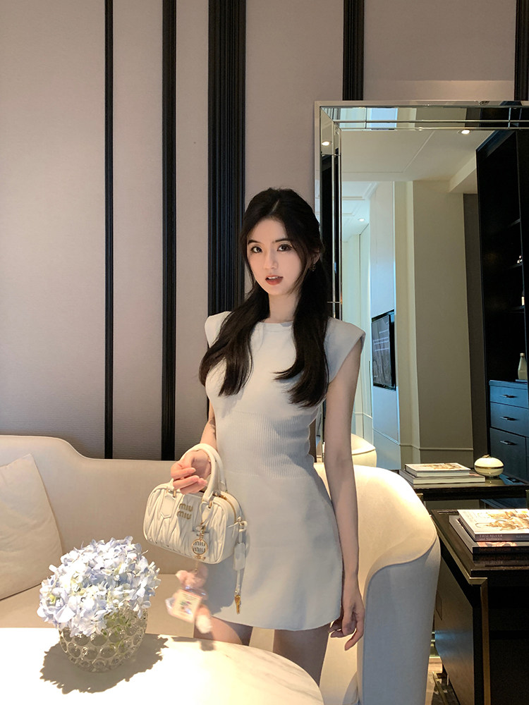 Woman wearing a white sleeveless dress and holding a white handbag in a modern interior setting.