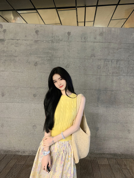 Woman wearing a yellow sleeveless knit top and floral skirt with a beige bag.