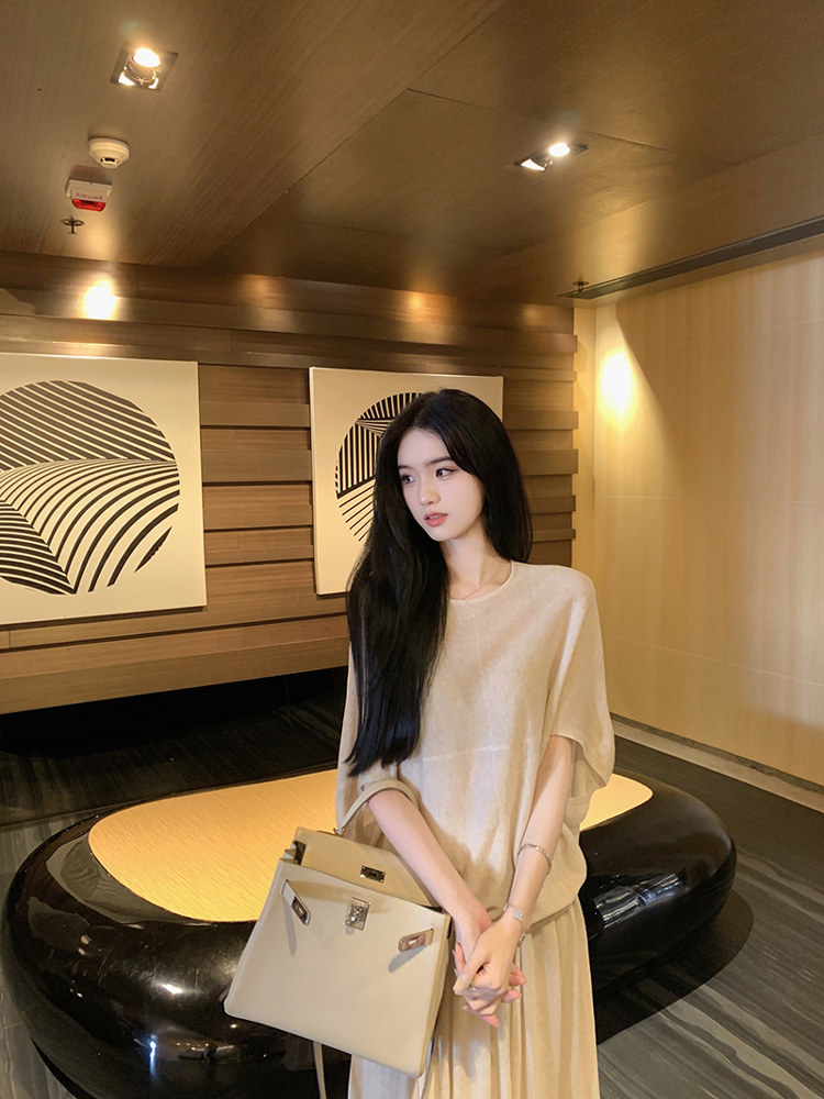 Woman wearing beige oversized top and matching skirt, holding beige handbag in modern interior