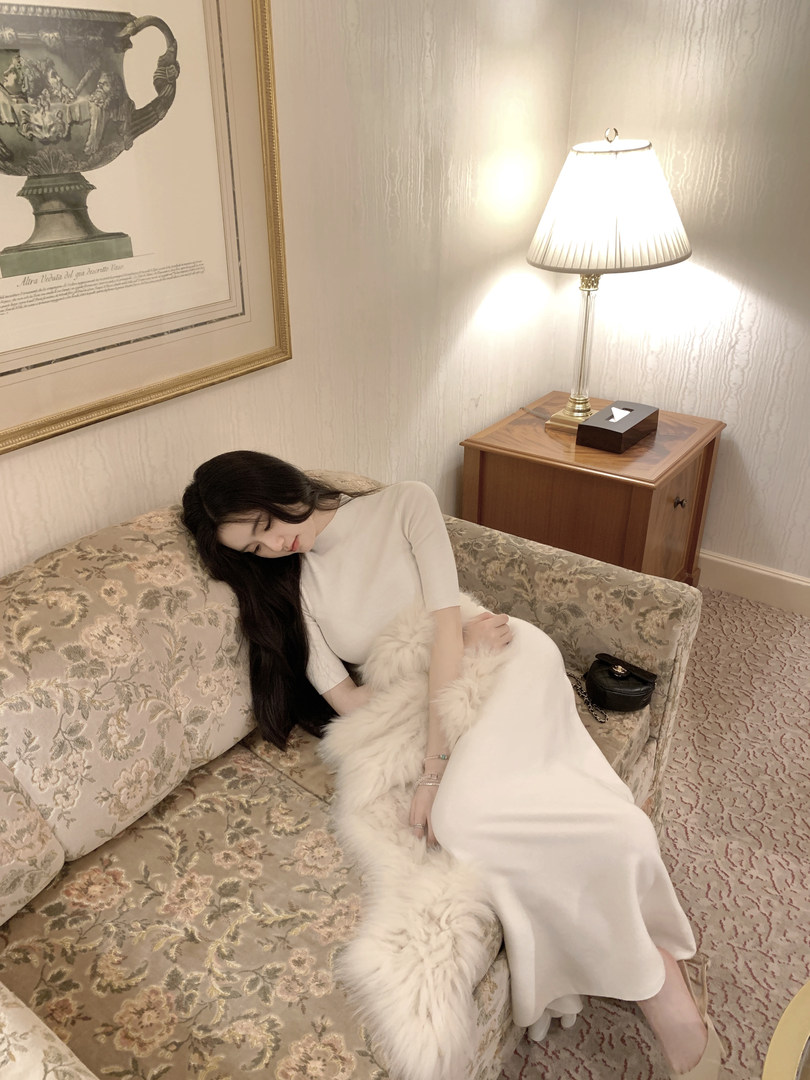 Woman wearing a white long-sleeve dress with a white faux fur stole, sitting on a floral sofa in a warmly lit room.
