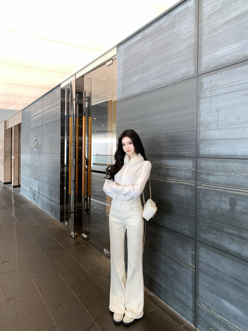 Woman wearing white knit cardigan with faux fur collar, matching wide-leg pants, and white quilted bag, standing against gray wall