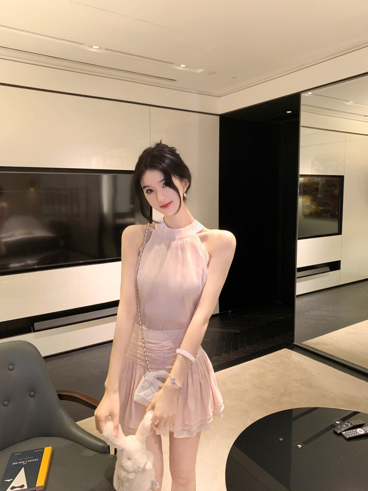 Woman wearing a light pink sleeveless dress with high neckline and pleated skirt, holding a small white bag and plush toy.