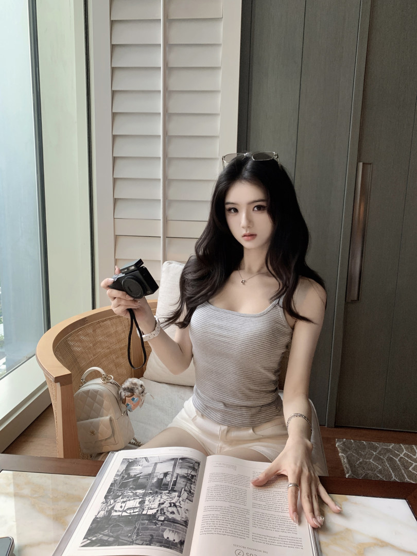 Woman wearing striped tank top and shorts, holding camera, seated by window with open book and handbag nearby
