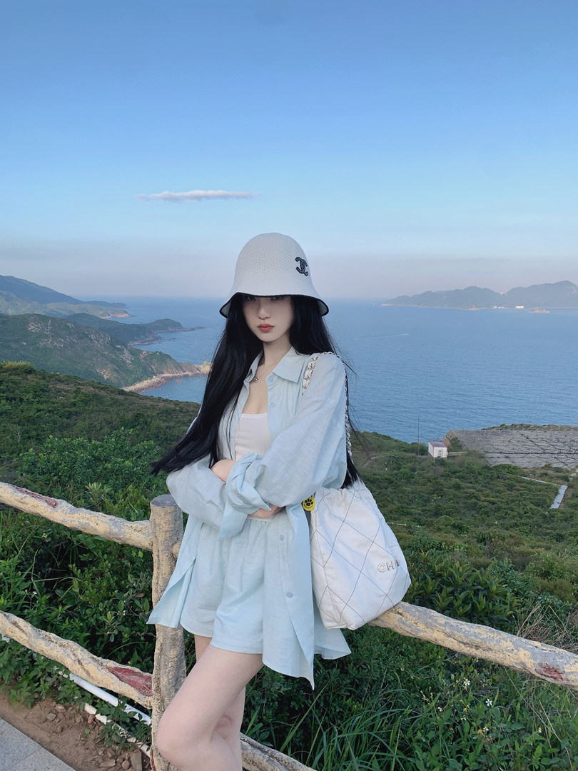 Woman wearing light blue shirt and shorts with white bucket hat and quilted bag, posing outdoors by the sea