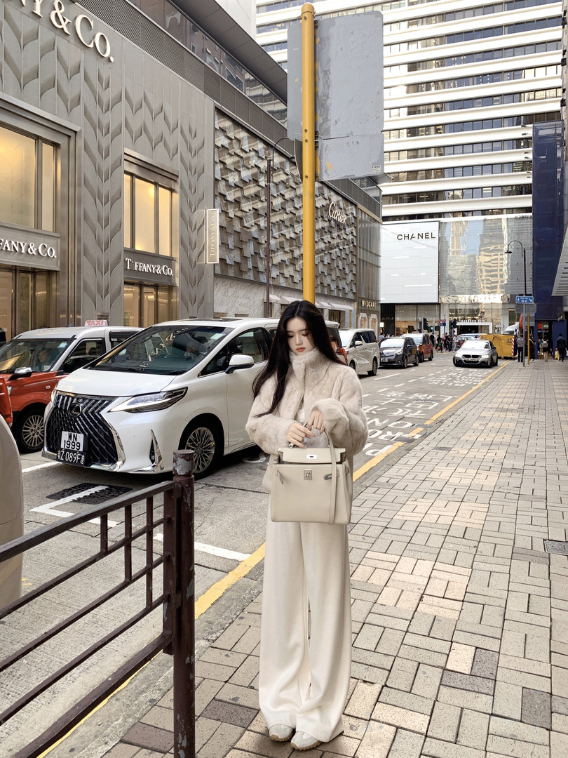 Woman wearing a beige fur coat and wide-leg pants, holding a beige handbag on a city street.