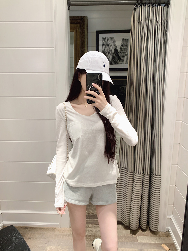 A person wearing a white long-sleeve top, gray shorts, and a white cap, taking a mirror selfie.