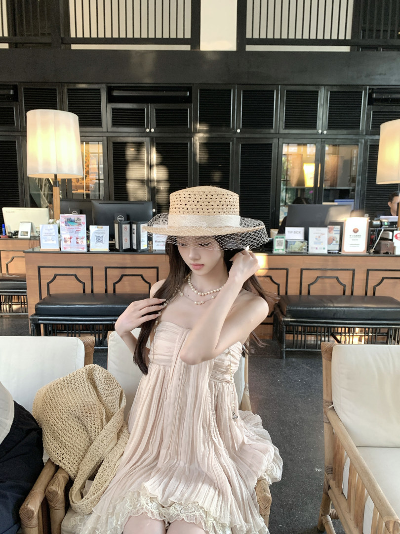 Woman wearing a light pink pleated dress and straw hat with veil, seated in a lounge setting