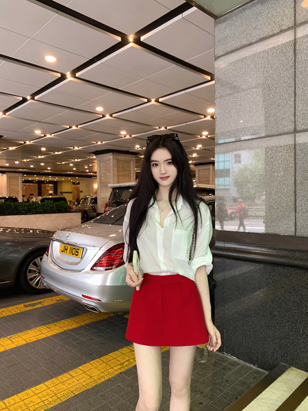 Woman wearing white button-up shirt and red mini skirt, standing outdoors near a car.