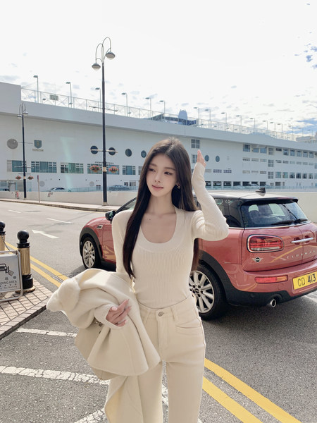 Woman wearing a cream ribbed top and matching pants, holding a coat, standing outdoors near a red car.