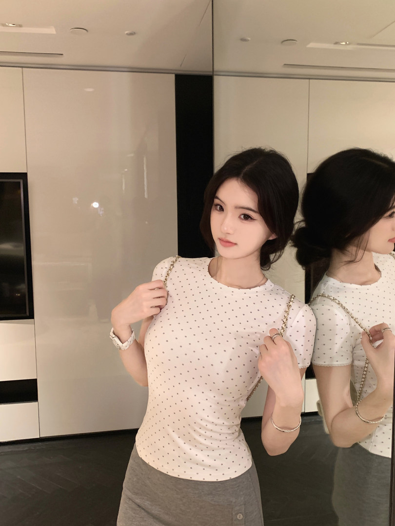 Woman wearing a white polka dot top with gray skirt, posing in front of a mirror.