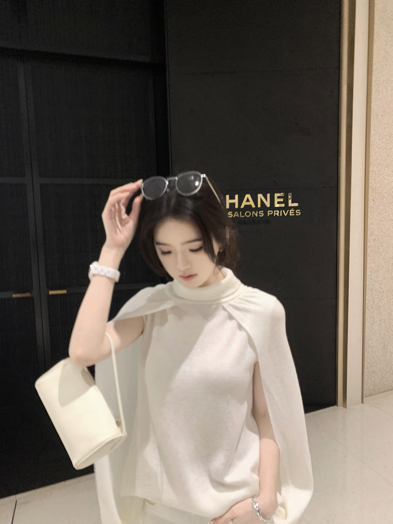Woman wearing white cape-style top with sunglasses on head, standing in front of Chanel store sign