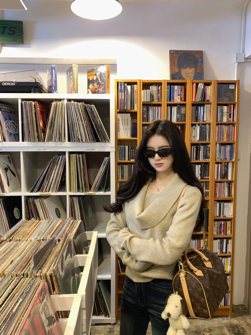 Woman wearing a beige ribbed sweater and sunglasses, standing in a record store with vinyl shelves and a Louis Vuitton bag.