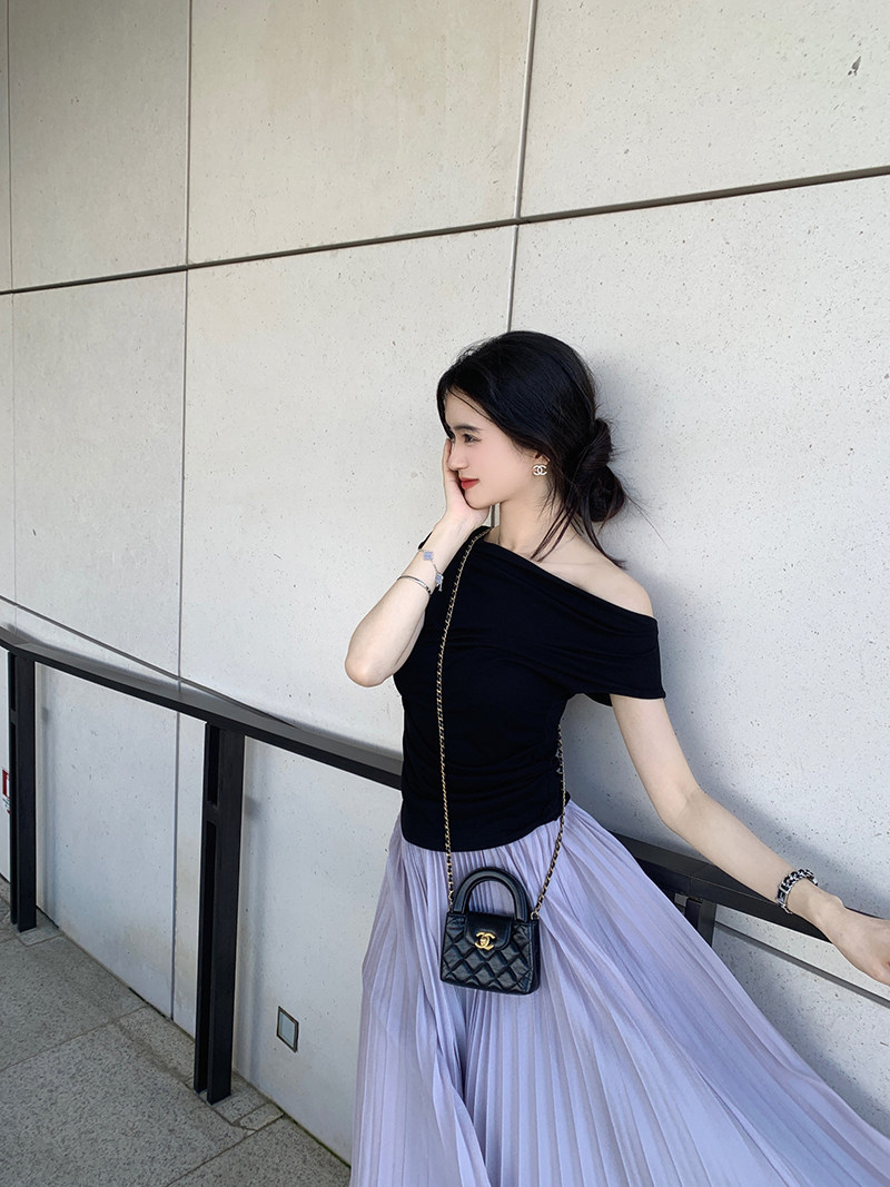 Woman wearing black off-shoulder top and lavender pleated skirt with a small black handbag.
